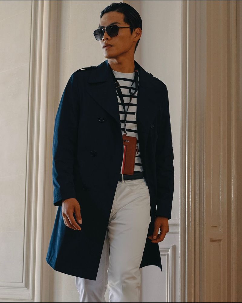Asian man in navy trench coat, striped shirt, and white pants stands indoors with sunglasses, looking to the side in a fashion shot.