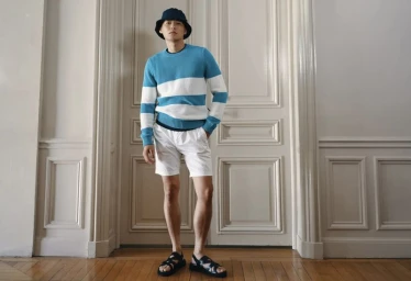 Model wearing a blue and white striped sweater, white shorts, bucket hat, and sandals standing against panelled double doors.