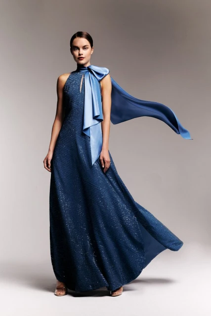 Full-length shot of a woman in an elegant blue gown with a large bow accent, posing in a studio setting with a soft, neutral backdrop.