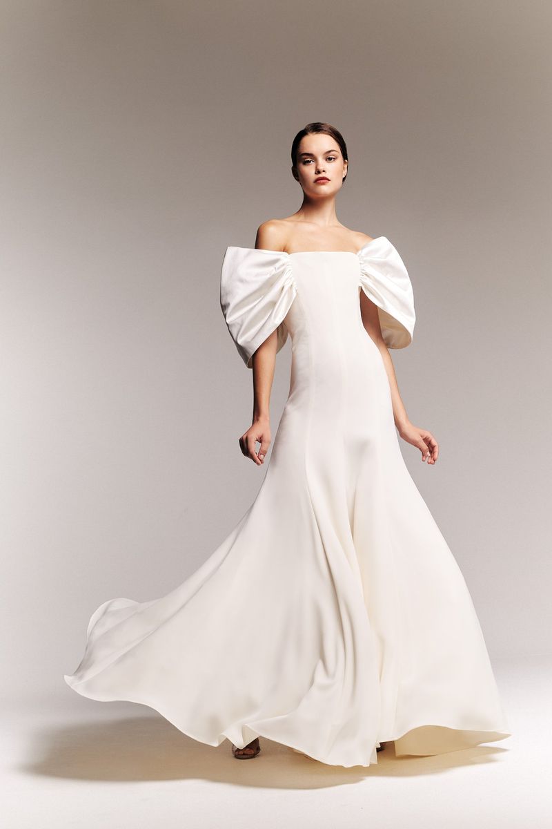 Woman in flowing white off-the-shoulder gown with large bow-like details, posing in a studio with soft lighting. Fashion editorial.