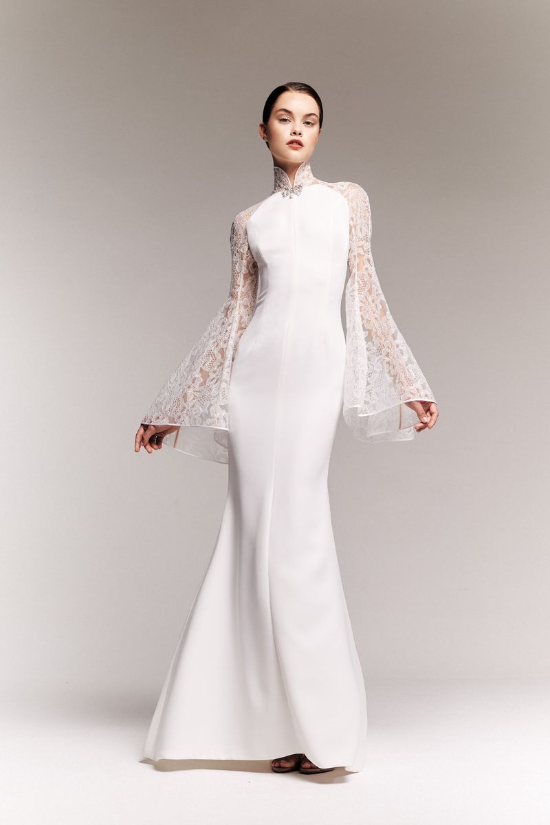 Fashion shot of a woman in a long white gown with lace sleeves, posing in a studio with a neutral background.