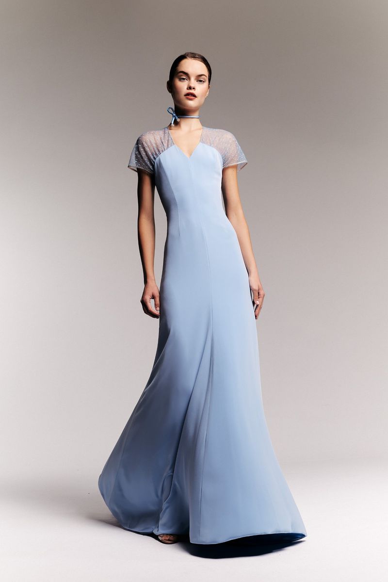A woman stands in a photo studio wearing a long periwinkle dress with a sheer dotted bodice. She poses in front of a graduated gray background.
