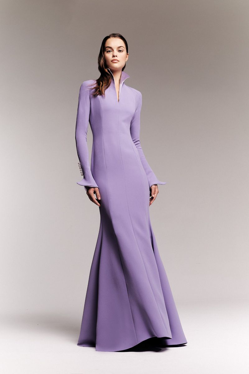 Elegant woman poses in a long-sleeved, floor-length lavender gown with a high collar in a fashion studio against a pale grey background.