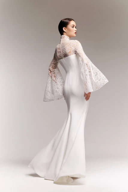 Elegant woman in a white lace-sleeved gown poses in a studio, showcasing high fashion and couture design with a focus on bridal style.