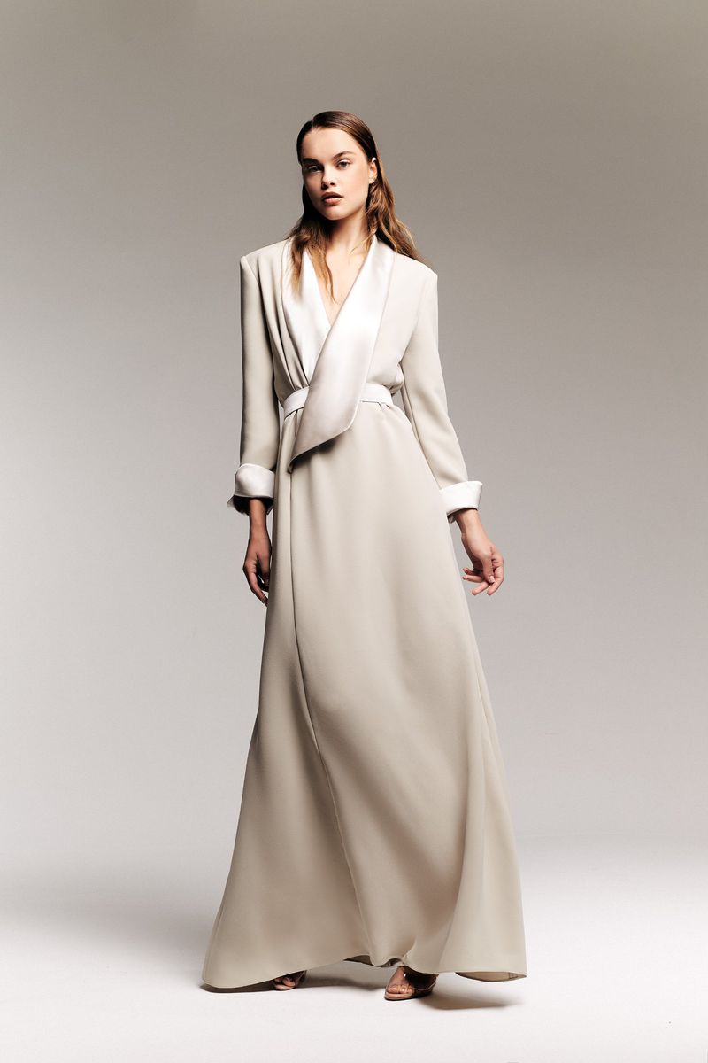 Full-length shot of a female fashion model in a light tan gown with white trim against a neutral grey studio backdrop, minimalist fashion.