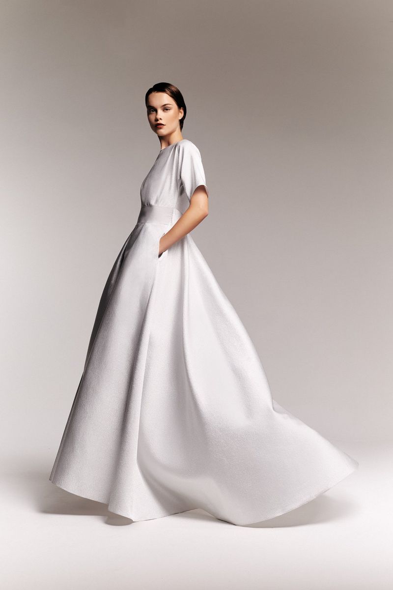 Elegant woman poses in a flowing, textured white gown with short sleeves and pockets against a soft grey backdrop in a fashion studio shot.