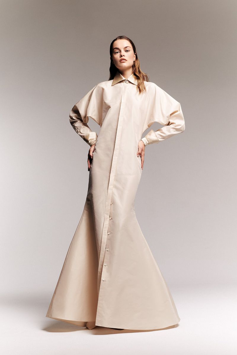 A tall, slender fashion model poses in a floor-length button-down dress in a bright studio setting with her hands on her hips.