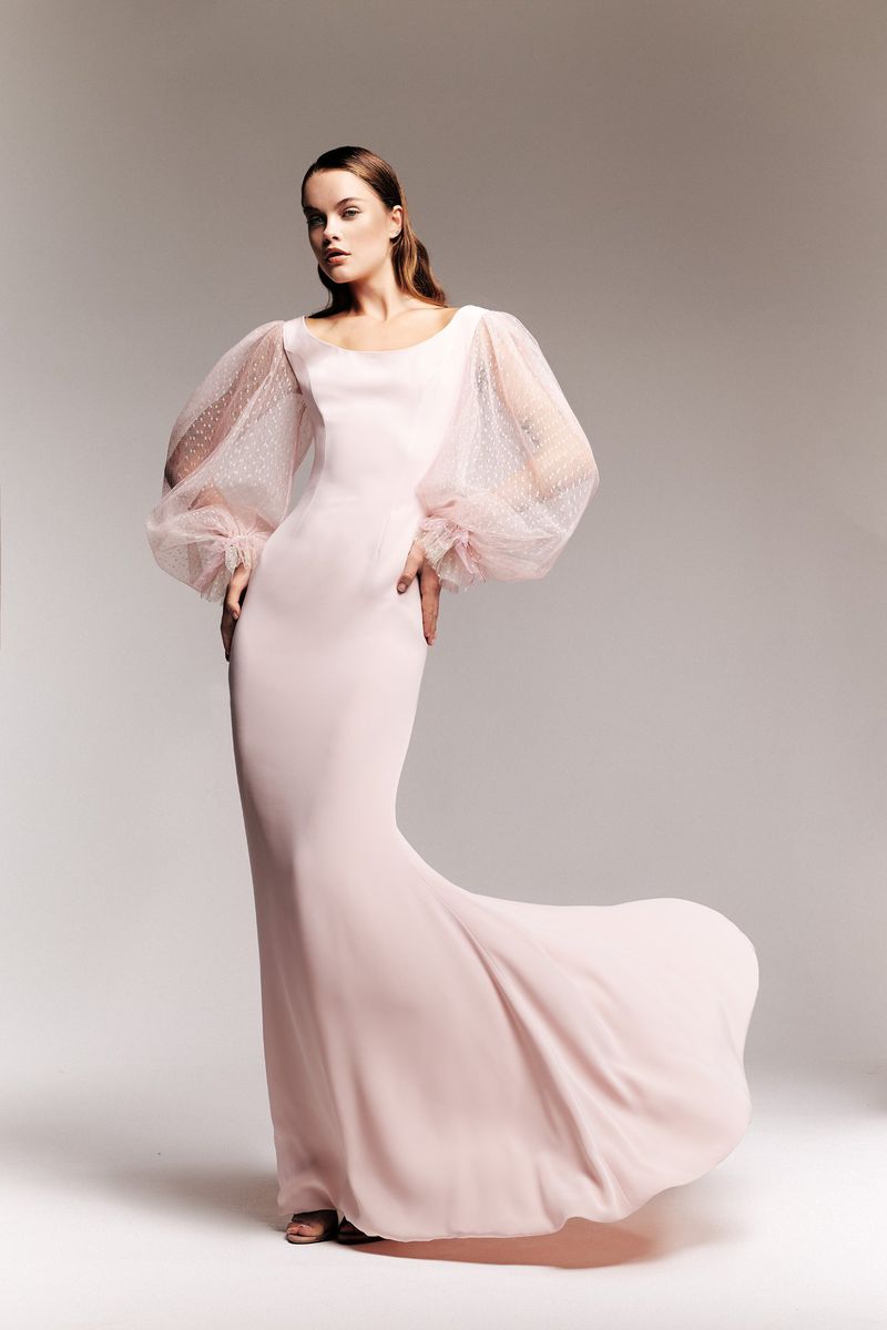 Elegant woman poses in long pink gown with flowing train and voluminous, sheer, dotted sleeves in a fashion studio setting.