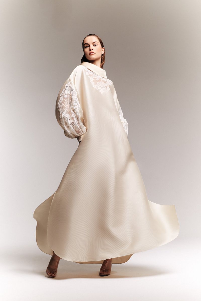 A woman in a flowing white dress with lace sleeves poses in a studio with an off-center composition, against a neutral gradient backdrop.