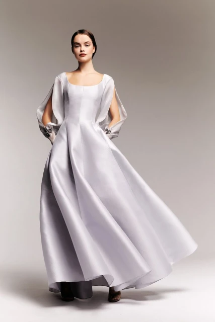 Elegant woman posing in a light grey gown with open sleeves, studio lighting, medium shot, fashion editorial photography.
