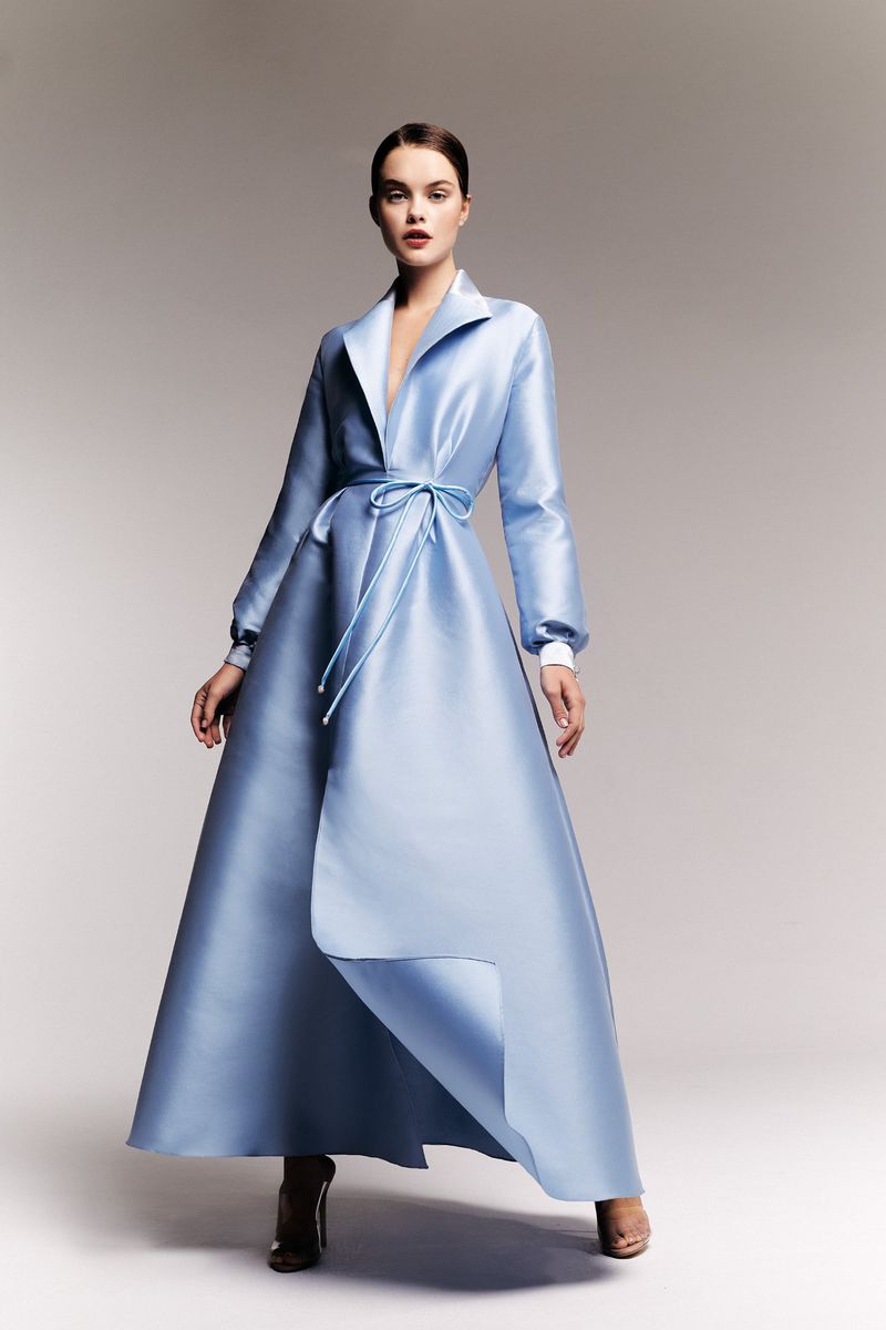 Elegant woman posing in a long, light blue satin coat dress with a tie waist in a studio setting with soft, neutral lighting.