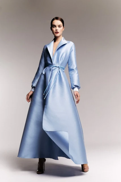 Elegant woman posing in a long, light blue satin coat dress with a tie waist in a studio setting with soft, neutral lighting.