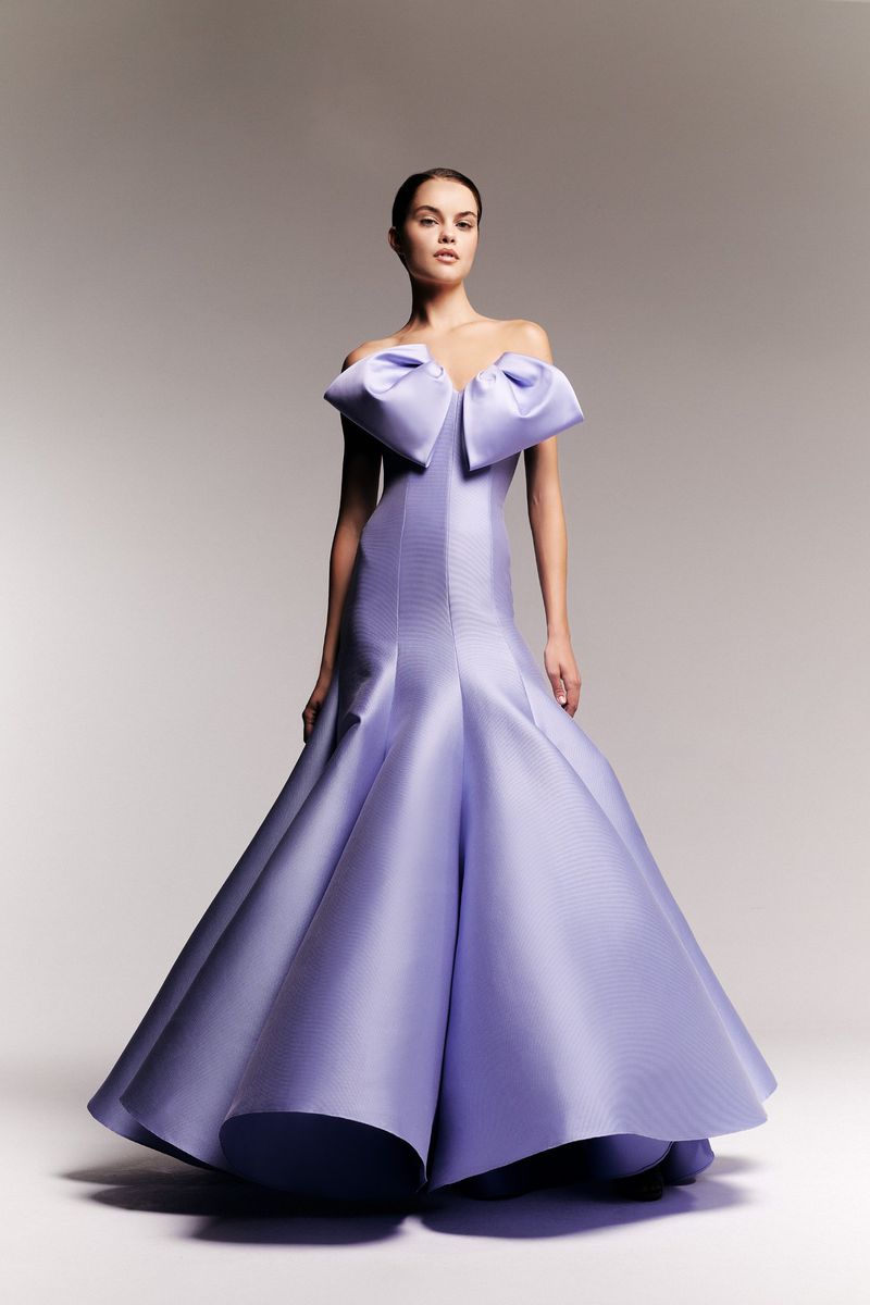 A woman wearing a lilac-colored, floor-length off-the-shoulder gown with a large bow detail poses in front of a light gray background.