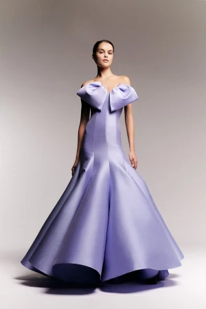 A woman wearing a lilac-colored, floor-length off-the-shoulder gown with a large bow detail poses in front of a light gray background.
