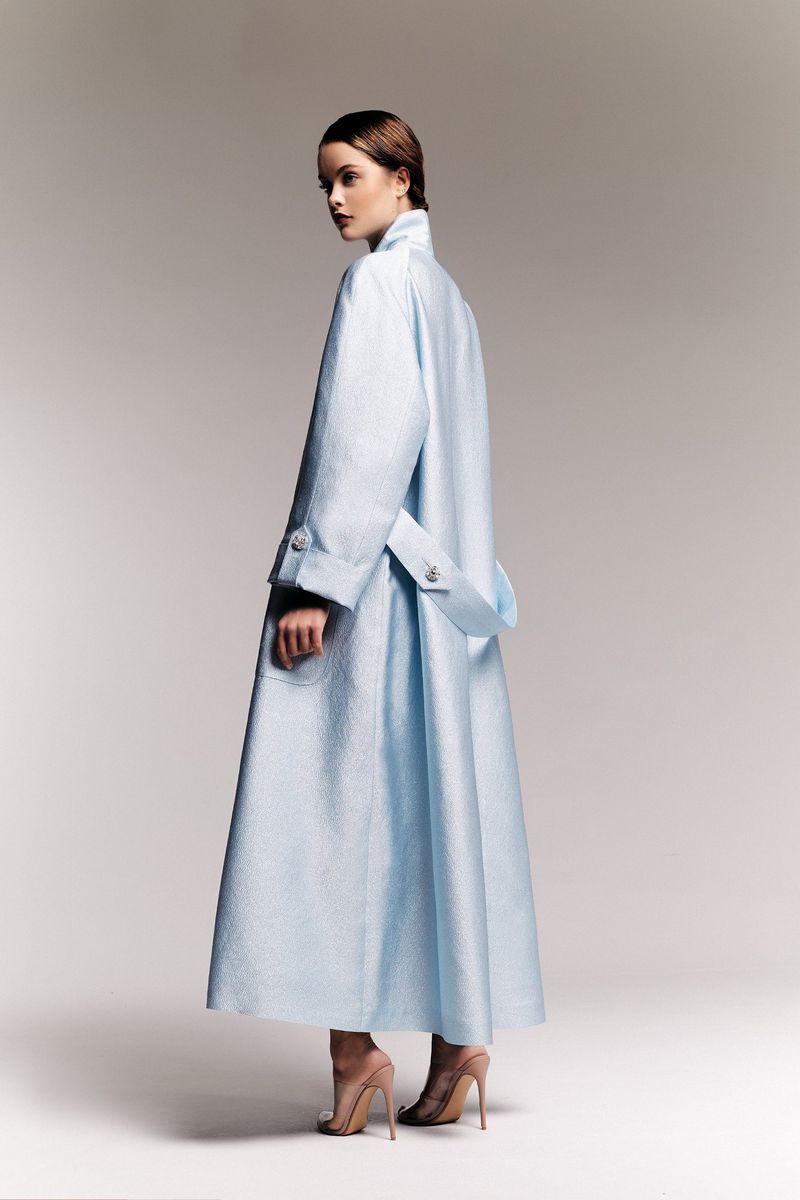 Fashion shot of a woman in a light blue coat, looking over her shoulder in a studio setting, showcasing modern elegance and style.