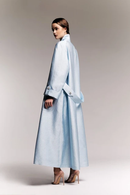 Fashion shot of a woman in a light blue coat, looking over her shoulder in a studio setting, showcasing modern elegance and style.