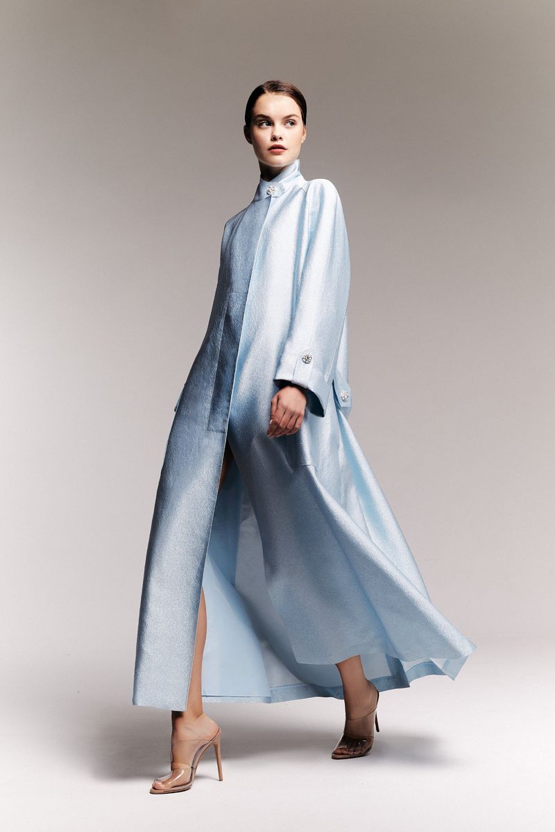 Full-length shot of an adult female fashion model posing in a long, light-blue coat dress and translucent high heels in a photo studio.