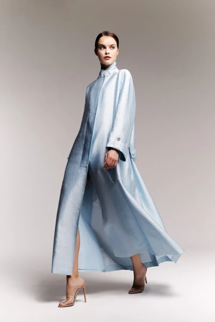 Full-length shot of an adult female fashion model posing in a long, light-blue coat dress and translucent high heels in a photo studio.