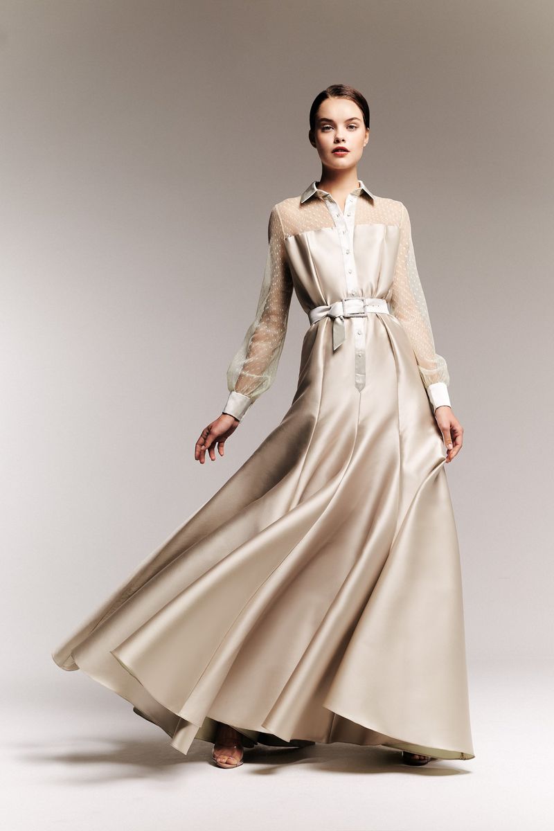 Elegant woman in a floor-length gown with sheer sleeves and a white belt, posing in a studio setting with soft lighting. Fashion editorial.