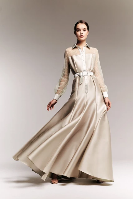 Elegant woman in a floor-length gown with sheer sleeves and a white belt, posing in a studio setting with soft lighting. Fashion editorial.
