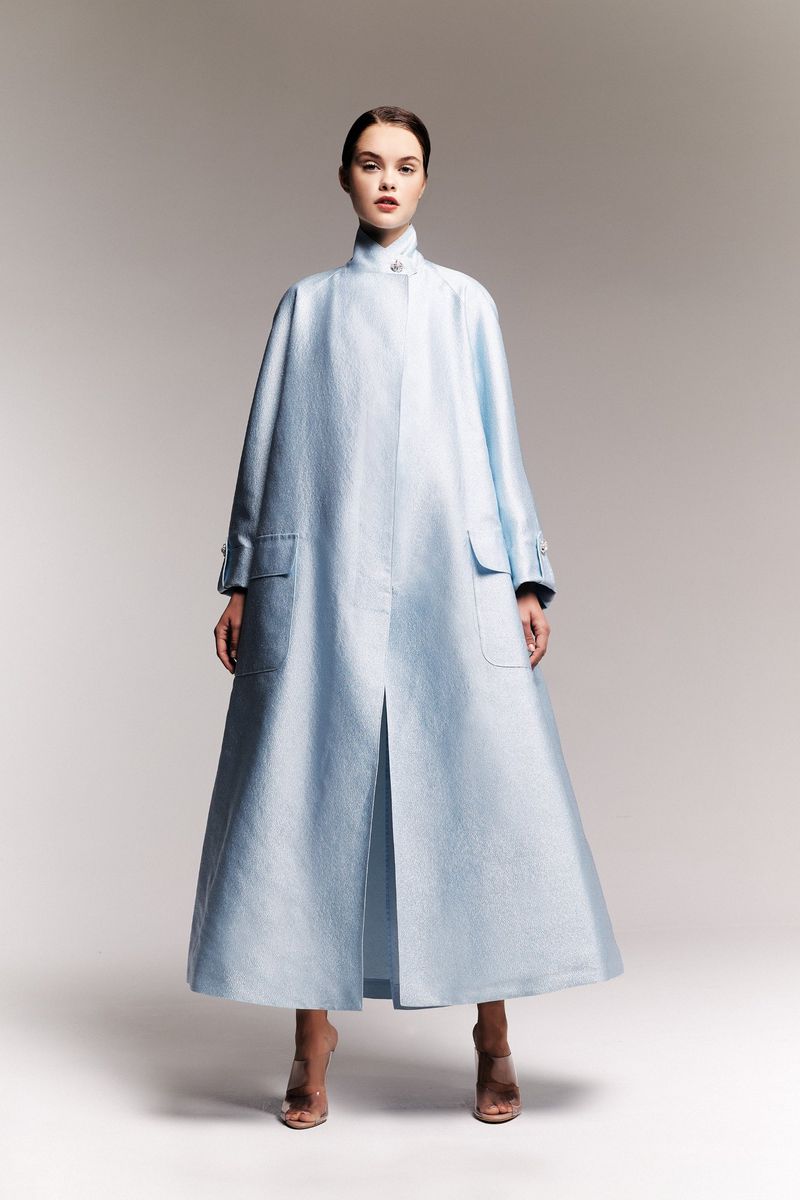 A woman posing in a light-blue, long-sleeved coat with pockets and nude high heels against a neutral background in a studio shot.