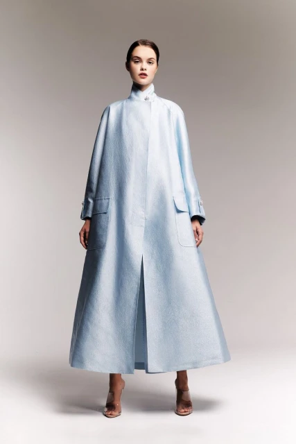 A woman posing in a light-blue, long-sleeved coat with pockets and nude high heels against a neutral background in a studio shot.
