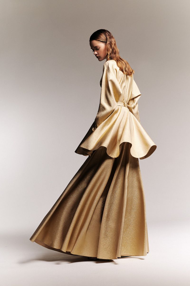 Model wearing long gold gown with an open back poses in a photo studio against a neutral background. Fashion editorial shot.