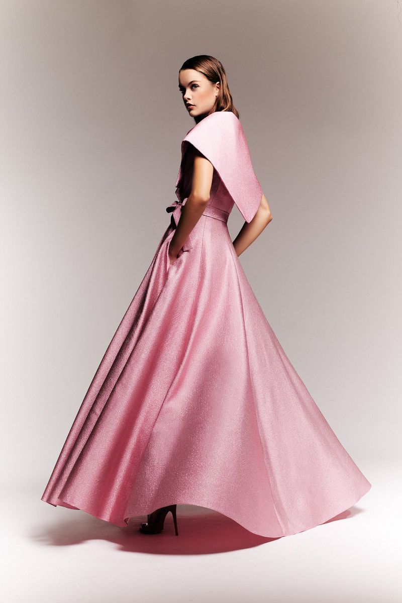 A woman poses in a formal pink gown with a dramatic shawl collar, in a studio setting with soft lighting.
