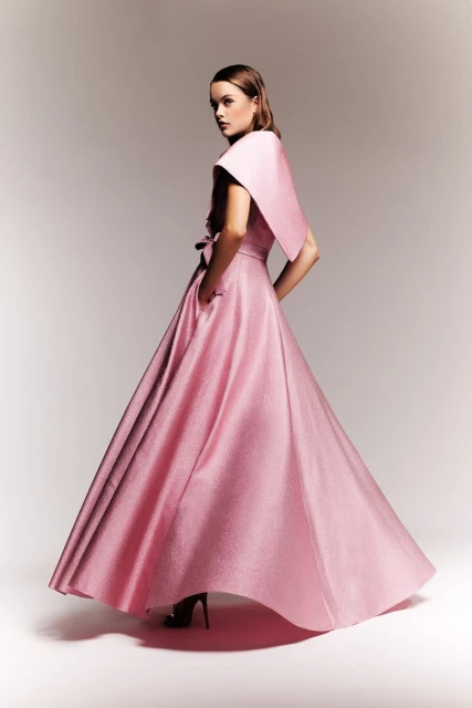 A woman poses in a formal pink gown with a dramatic shawl collar, in a studio setting with soft lighting.