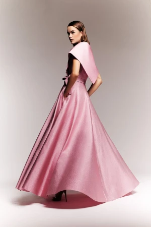 A woman poses in a formal pink gown with a dramatic shawl collar, in a studio setting with soft lighting.