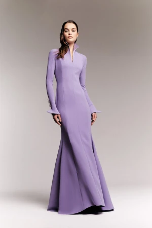 Model wearing a long lavender dress with a high collar standing against a light grey studio background.