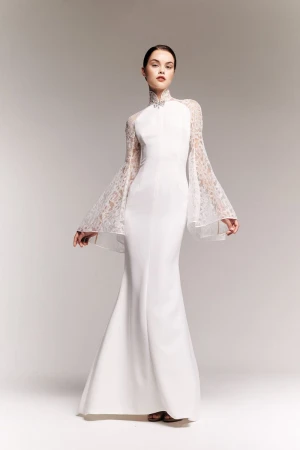 Model wearing a white gown with sheer lace bell sleeves and a high collar against a neutral studio backdrop