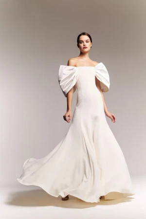 Model wearing a white off-the-shoulder gown with dramatic sleeves against a gradient studio background.