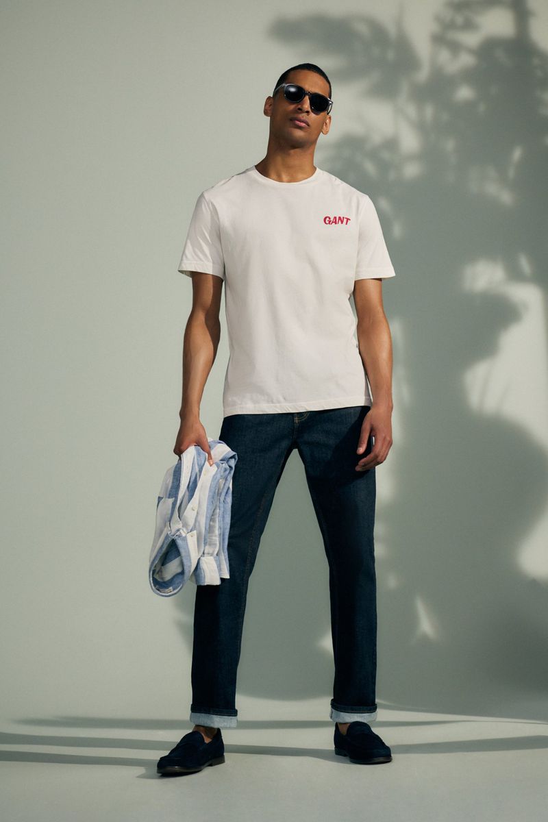 Male model in t-shirt and jeans poses in a studio with soft lighting and a subtle palm tree shadow. He's holding a shirt in hand.