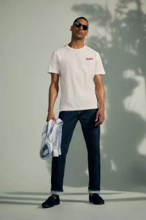 Man wearing a white t-shirt and dark jeans standing against a light grey seamless backdrop with leaf shadow patterns.