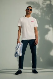 Man wearing a white t-shirt and dark jeans standing against a light grey seamless backdrop with leaf shadow patterns.