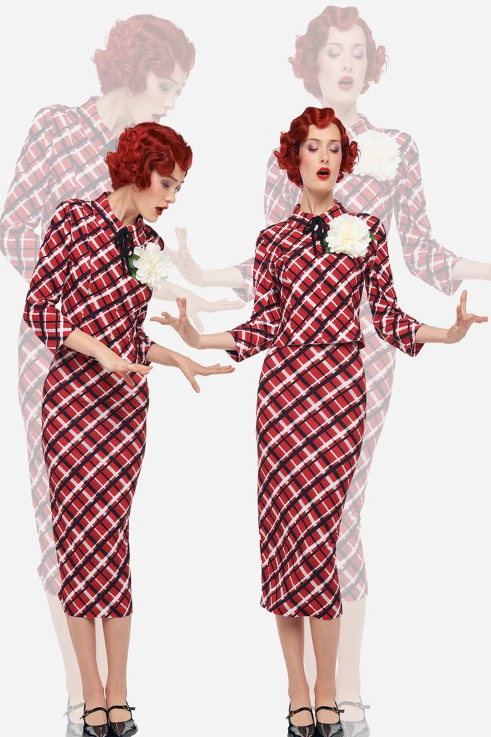 Model with red hair wearing a red plaid dress with a white floral accent against a plain white background