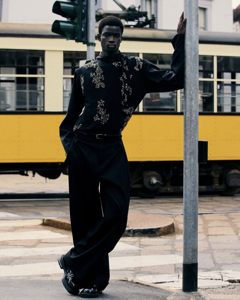 A dark-skinned male model in an embellished black outfit poses in front of a yellow tram and a traffic light on a city street.
