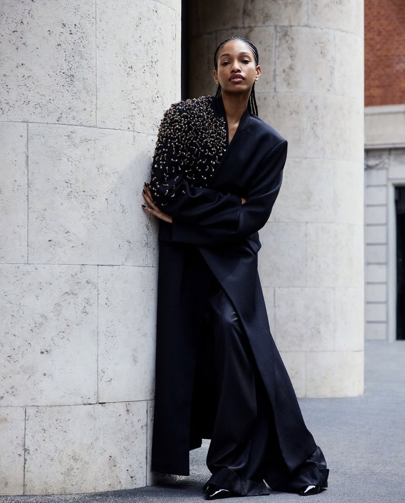 A model leaning against a stone pillar outdoors wearing a black embellished coat for Emporio Armani Fall Winter 2024/25.