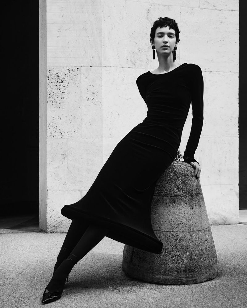 A stylish young woman in a long black dress leans against a concrete pillar, striking a pose in an outdoor fashion shot.