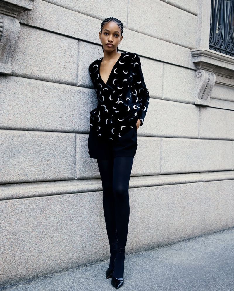 Fashionable woman in black moon-patterned jacket, black shorts, and tights posing against a textured gray urban wall backdrop.