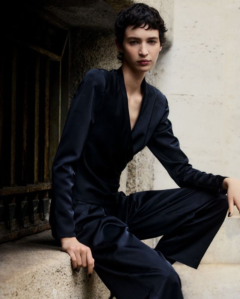 A woman in a navy satin suit sits on steps, posing for a fashion shot in natural light. The background is an urban stone wall.