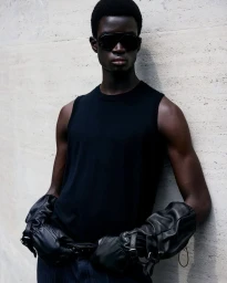 Black man wearing dark sunglasses, black sleeveless top, and leather gloves leaning against a light stone wall.
