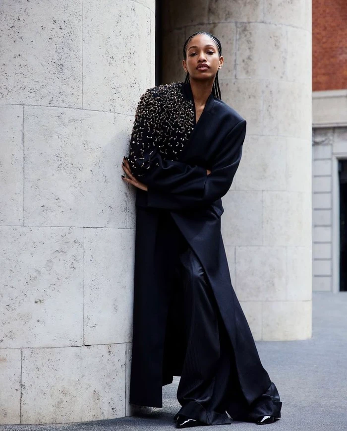 A model leaning against a stone pillar outdoors wearing a black embellished coat for Emporio Armani Fall Winter 2024/25.
