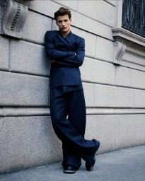 A young man wearing a dark blue suit leaning against a stone building wall with crossed arms.