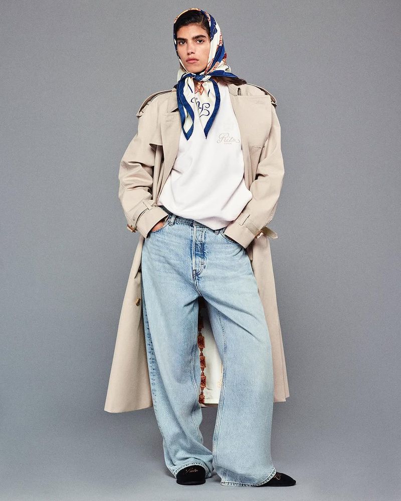 A woman poses in wide-leg jeans, a white sweatshirt, and a headscarf against a grey backdrop, her hands casually in her pockets.