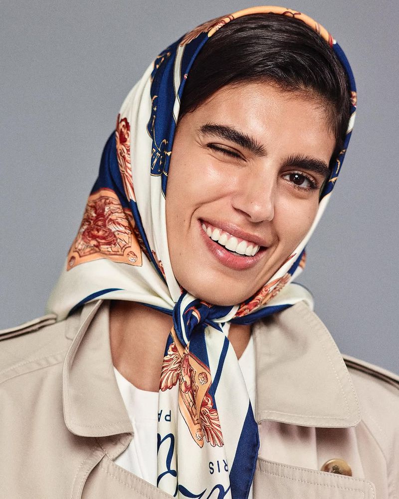 Smiling woman wearing a patterned headscarf and trench coat, winking at the camera against a gray background, close-up studio portrait.