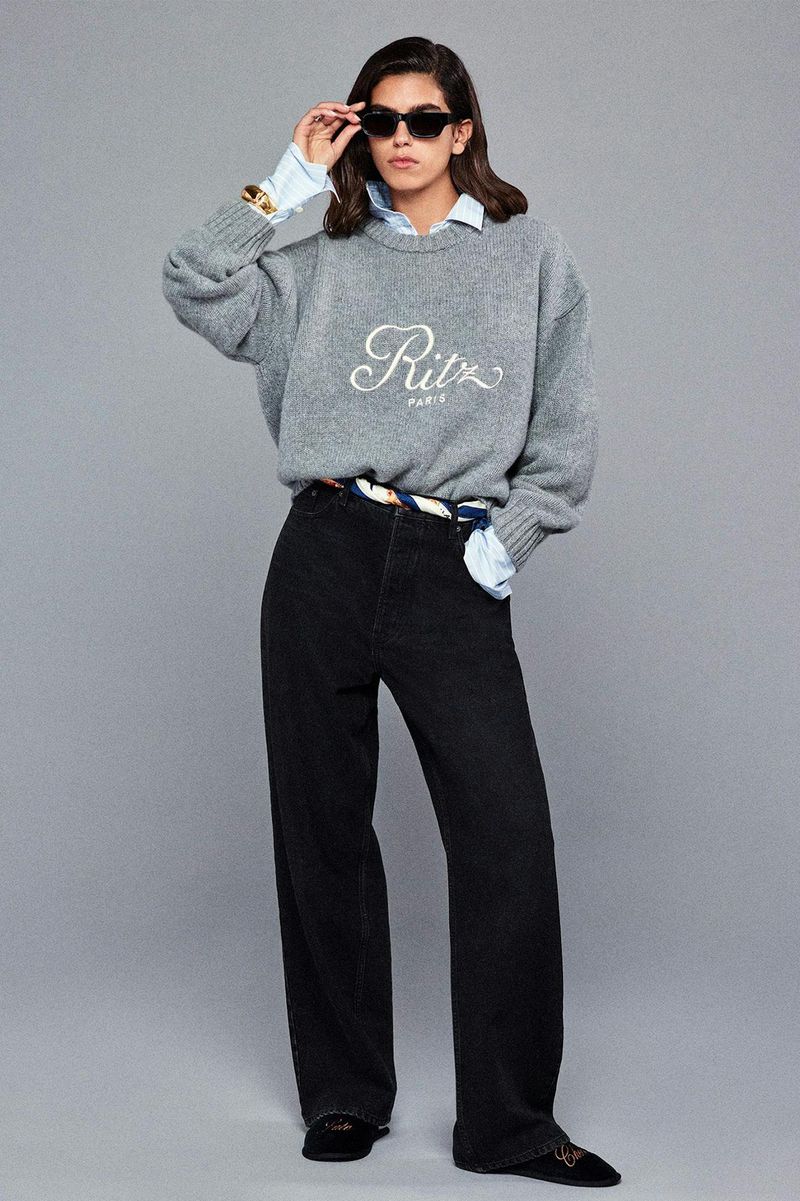 A woman in sunglasses, a gray sweater with 'Ritz Paris' in script, black jeans, and black shoes poses against a gray backdrop.