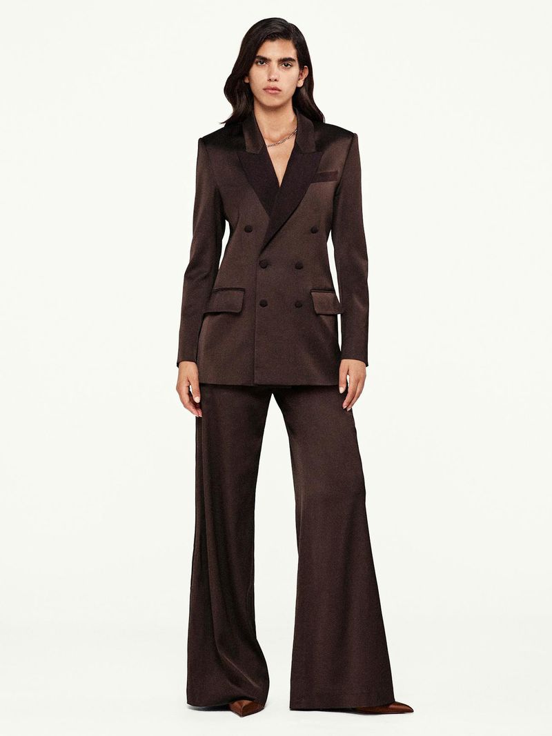 A woman in a satin suit poses in front of a white background in a full shot. The suit is a chocolate brown color with dark lapels.