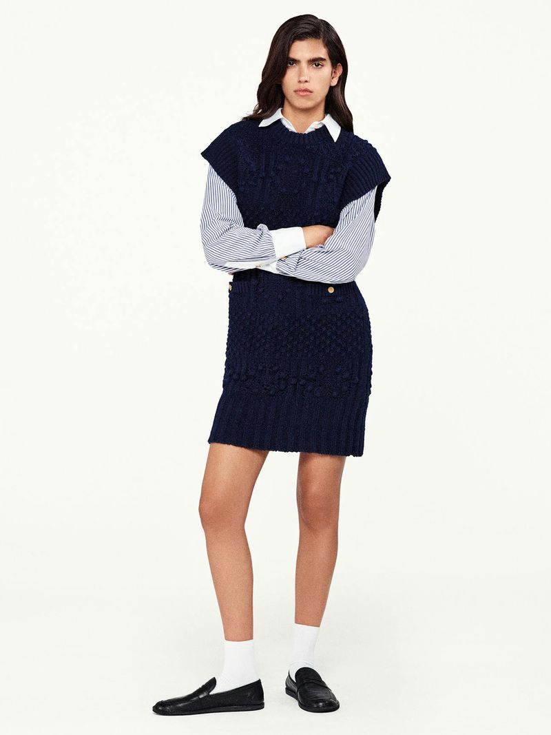 Woman stands in a photo studio wearing a navy cable knit sweater vest and matching skirt, striped shirt, white socks, and black loafers.